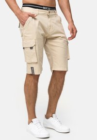 Shorts cargo beiges avec une coupe droite, longueur genou, plusieurs poches, et une ceinture noire avec logo. Accompagnés de baskets blanches.