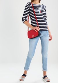 Röd axelväska med marinblå detaljer, slät yta och rund form. Modell som bär en marinblå och vit randig skjorta, ljusblå jeans och silverfärgade sandaler.