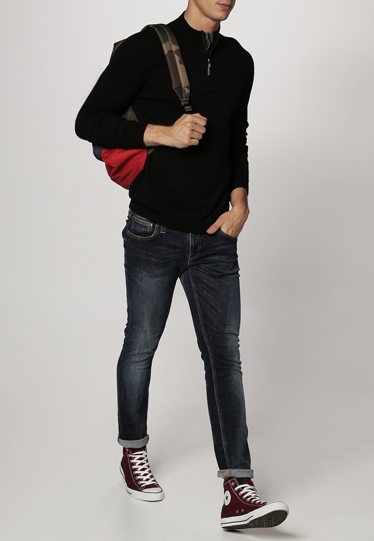 Jeune homme portant un pull noir, un jean foncé, des baskets montantes bordeaux, portant un sac à dos rouge et camouflage sur une épaule.