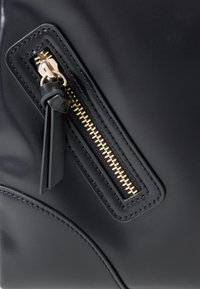 Material sintético negro con un acabado liso, que presenta una cremallera dorada y un tirador de cremallera de cuero. La costura de contorno añade textura.