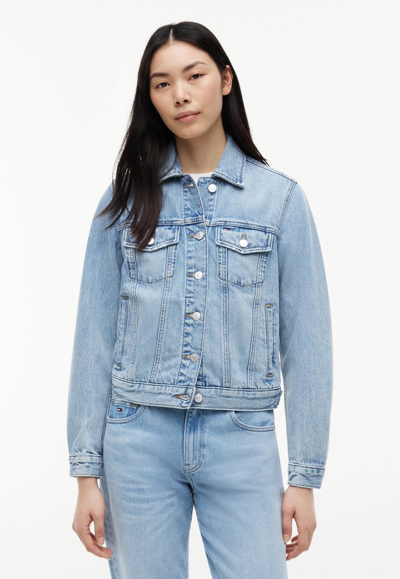Femme aux longs cheveux foncés portant une veste en jean bleu clair et un jean assorti, debout devant un fond blanc uni.