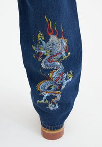 Džínové kalhoty s multicolor designem draka vytvořeným z kamínků, s podrobnými vzory v odstínech modré, červené a žluté.