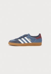 Zapatillas de gamuza azul con franjas blancas y el logo dorado "Gazelle". Presenta una suela de goma y acentos en color navy en el cuello y la lengüeta.