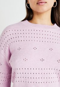 Pulover tricotat roz deschis, cu guler rotund și un model de mici orificii pe întreaga suprafață pentru un plus de textură. Mâneci lungi.