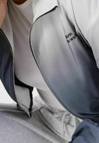 Giacca a zip in gradiente grigio e blu con colletto alto, dotata di hardware argentato e piccolo dettaglio del logo, abbinata a una camicia bianca.