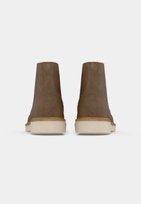 Paio di stivaletti alla caviglia in suede marrone con suole in gomma spessa beige chiaro, visti da dietro su uno sfondo bianco.