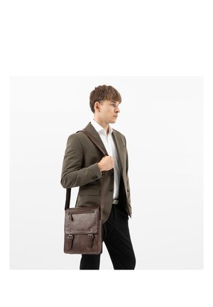 Jonge man in een bruine blazer en wit overhemd draagt een donkerbruine leren schoudertas, staande tegen een witte achtergrond.
