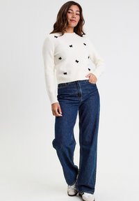 Pull en tricot blanc avec des embellissements en forme de nœud noir, col rond et manches longues, associé à un jean en denim bleu taille haute et des baskets.