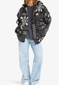 Teddy svart och vit mönstrad fleecejacka med blommiga och geometriska mönster, med huva och knappdetalj, ihop med ljusblå jeans.