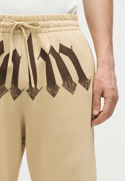 Μπεζ παντελόνι φόρμας με μαύρο γοτθικό σχέδιο μπροστά και σούρα στη μέση, φορεμένο με λευκό πουκάμισο και χέρι ακουμπισμένο δίπλα.