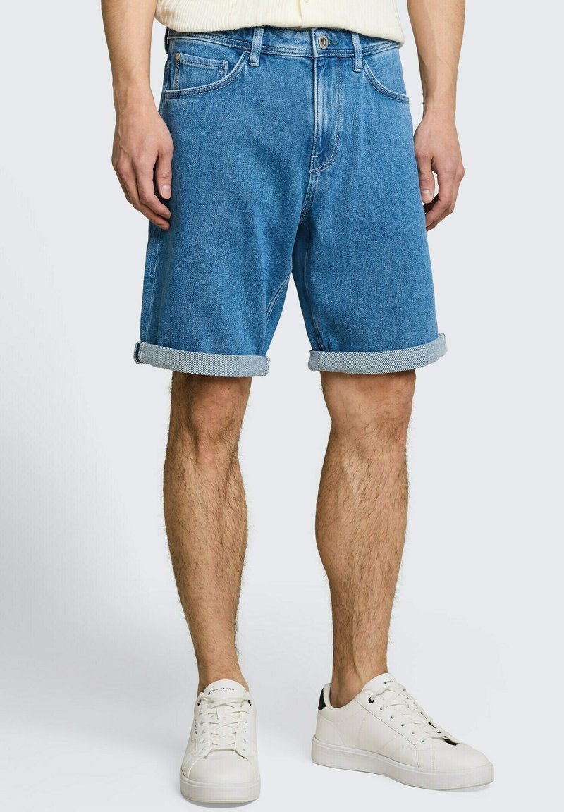 Homme portant un short en jean bleu retroussé, des baskets blanches et une chemise de couleur claire, les mains détendues le long du corps.
