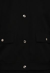 Jachetă neagră cu două buzunare frontale, nasturi capsa argintii, țesătură texturată și margine cu dungi elastice. Potrivită pentru purtarea casual.