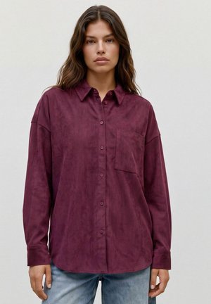 Jeune femme portant une chemise bordeaux ample à manches longues avec une poche poitrine et un jean bleu clair.