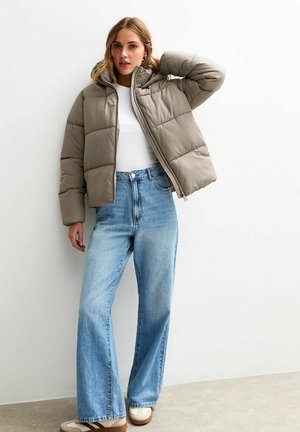 Beige gewatteerde jas met een capuchon, ritsluiting, gecombineerd met lichtblauwe wijde jeans en witte sneakers met bruine accenten. Gladde textuur.