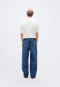 Λευκό πουκάμισο με κοντά μανίκια και άνετο μπλε τζιν. Το τζιν έχει δύο πίσω τσέπες και ένα καφέ δερμάτινο ταμπελάκι στη μέση.