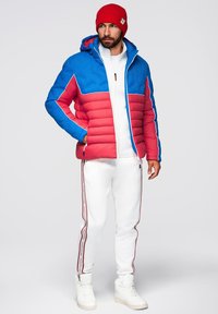 Vadderad jacka i blått och rött med dragkedja och huva. Kombineras med vita sweatpants med röda sidostrick och vita sneakers.