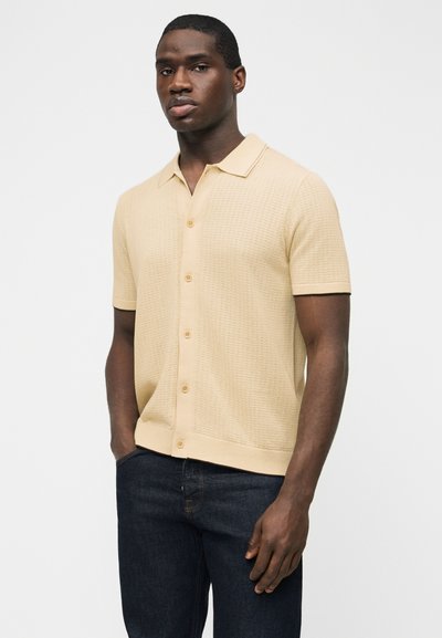 Άνδρας που φοράει πλεκτό πουκάμισο μπεζ με κοντό μανίκι και γιακά, συνδυασμένο με σκουρό μπλε τζιν, στέκεται μπροστά σε απλό φωτεινό φόντο.