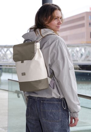 Jonge vrouw in grijze jas en spijkerbroek staat bij het water, draagt een beige en olijfgroene rugzak, kijkt over haar schouder.