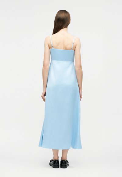 Γυναίκα με μακριά καστανά μαλλιά, γυρισμένη πλάτη, φοράει αμάνικο μίντι φόρεμα σε ανοιχτό μπλε χρώμα και μαύρα παπούτσια σε απλό φόντο.