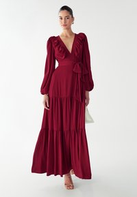 Dlhé, tmavočervené maxi šaty s volánovým výstrihom do v a dlhými pufovanými rukávmi a opaskom na zaväzovanie v páse. Sukňa má vrstvené vrstvy.