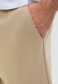 Pantalons beiges avec une texture lisse, poche intégrée, et design à jambe droite, partiellement obscurcis par une main.