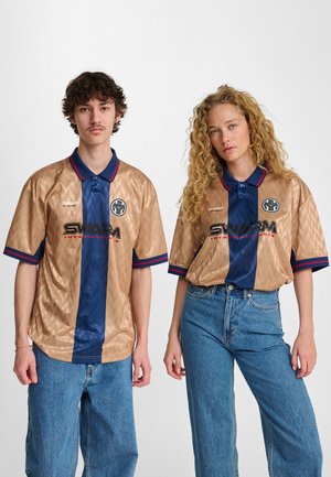 Koszulka z krótkim rękawem w kolorze brązowym i granatowym, z wzorzystą fakturą, niebieskim kołnierzem, czerwonymi akcentami i logo. Noszona z jasnoniebieskimi spodniami o szerokich nogawkach.