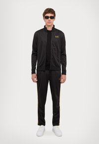 Black athletic jacket and pants set made of smooth fabric, featuring gold accents and a logo. Worn with white sneakers and dark sunglasses.