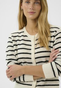 Jeune femme aux longs cheveux blonds, portant un cardigan blanc à rayures horizontales noires, bras croisés, expression neutre.