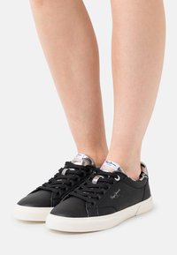 Mustat nahkakengät valkoisilla kumipohjilla, joissa on hopeanvärisiä yksityiskohtia kantapäässä, mustat nauhat ja teksturoitu pinnan viimeistely.