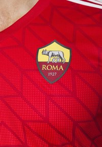 Maillot de sport rouge avec des motifs géométriques, arborant le blason du club de football AS Roma représentant la louve et les jumeaux avec le texte "ROMA 1927".