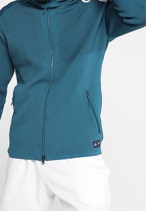 Person iført en teal lynlåstrøje med lynlåslommer og hvide bukser, stående mod en ensfarvet hvid baggrund.