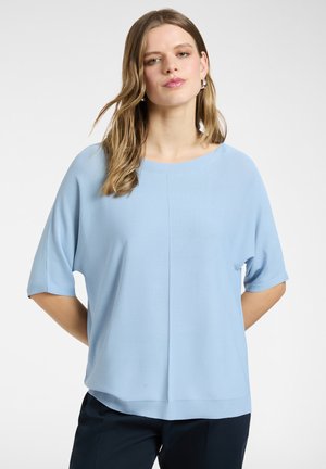 Młoda kobieta z długimi włosami, ubrana w luźną, jasno niebieską bluzkę z krótkim rękawem i ciemne spodnie, stojąca na jednolitym tle.