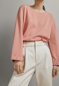 Light pink cotton long-sleeve top with a wide neckline, featuring studded accents on the cuffs. Paired with high-waisted white trousers.