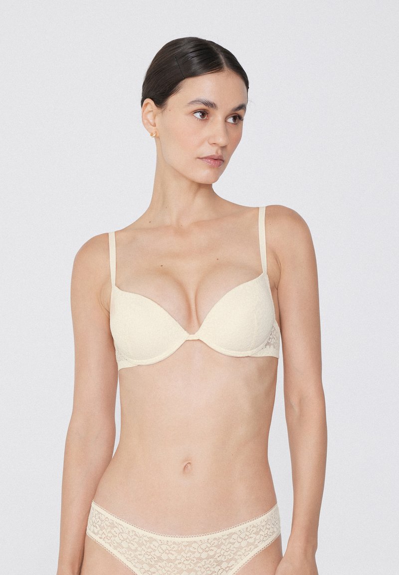Beige Spitzen-BH mit vorgeformten Cups und verstellbaren Trägern, der mit floralem Muster und nahtlosem Design für zusätzlichen Komfort versehen ist.