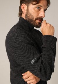 Man met baard, dragend van een zwarte gestructureerde coltrui, leunend met zijn kin op zijn rechterhand, iets naar rechts kijkend tegen een effen achtergrond.
