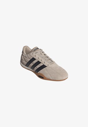 Zapatilla deportiva de ante beige con rayas negras, cordones y una suela de goma color chicle, mostrada en ángulo para mostrar detalles superiores y laterales exteriores.