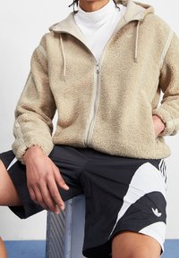 Fleece zip-up jacka i beige med mjuk textur, med hög krage och ljusgrå detaljer. Svarta shorts med vita och svarta designelement.
