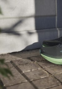 Scarpe da ginnastica grigie con superficie strutturata e intersuola verde, posizionate su una superficie di mattoni, parzialmente ombreggiate da foglie.
