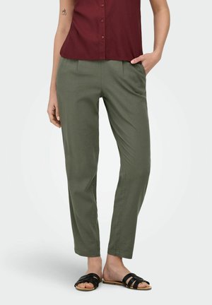 Kvinde iført olivengrønne bukser, bordeaux knapbundet skjorte, sorte sandaler, med den ene hånd i lommen og en lille tatovering på armen synlig.