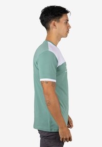 Camisa de manga corta en tela verde con un panel blanco en los hombros. Presenta un cuello redondo y una textura lisa. Vista lateral mostrada.