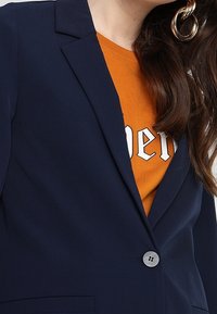 Femme portant un blazer bleu marine sur une chemise orange avec un texte blanc, cheveux longs bruns visibles, et une grande boucle d'oreille torsadée en or.