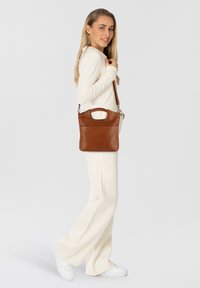 Borsa a tracolla in pelle marrone con manico superiore, tasca frontale e tracolla regolabile, caratterizzata da una texture liscia e un design minimalista.