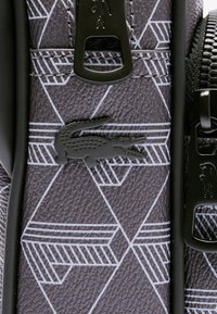 Bolsa sintética en negro y gris con patrones geométricos en blanco, que presenta un logo de cocodrilo negro elevado y detalles en hardware negro.