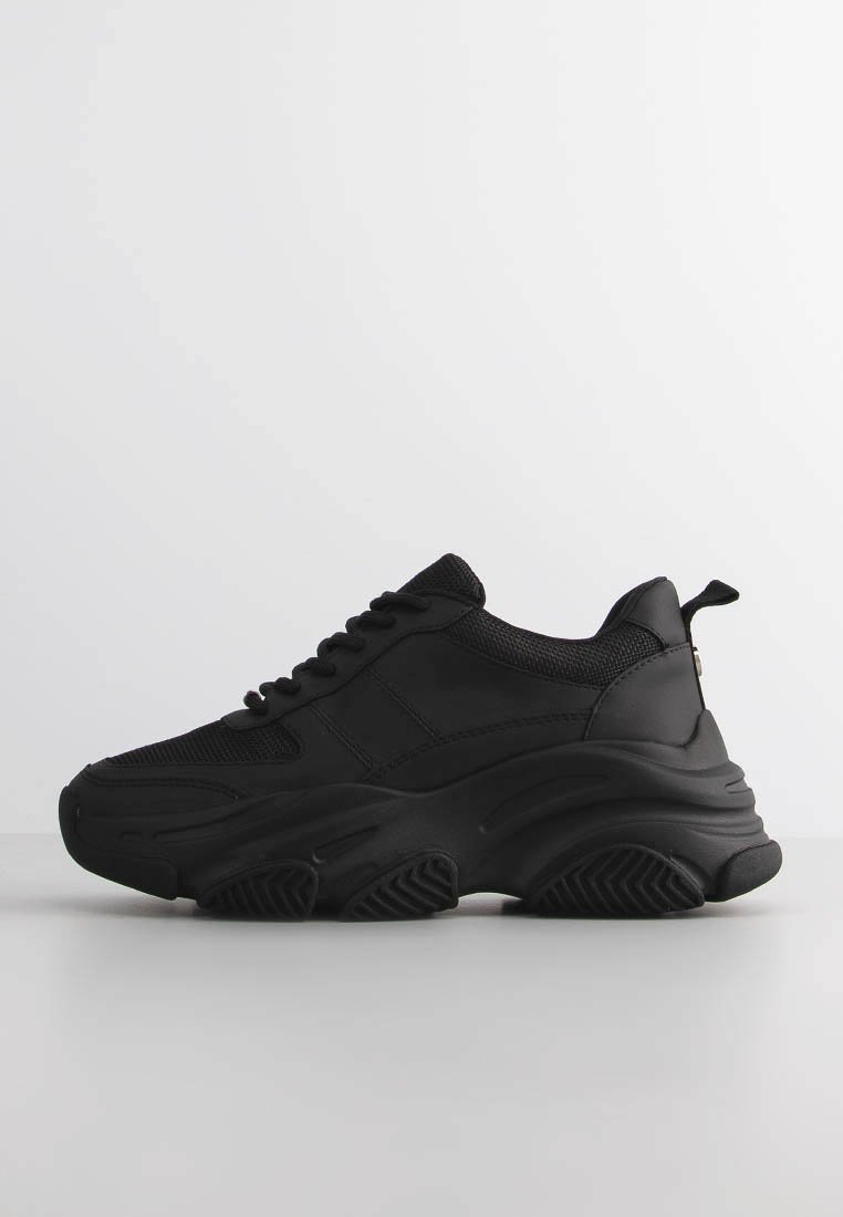 Svart sneaker med kraftig sula, detaljer i mesh och läder, snörningsdesign och dragflik på klacken, visad mot en helt vit bakgrund.