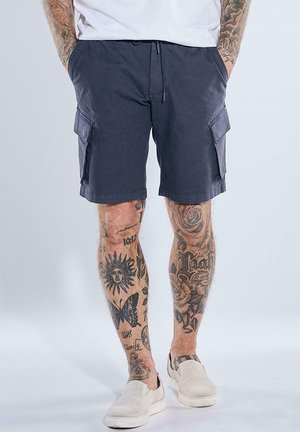 Mann trägt marineblaue Cargoshorts und weiße Slip-On-Schuhe, mit tätowierten Beinen und Händen in den Taschen vor einem einfarbigen hellen Hintergrund.