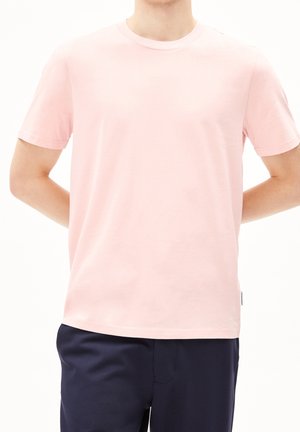 Osoba ubrana w prosty jasnoróżowy t-shirt z krótkim rękawem i ciemnoniebieskie spodnie, stojąca z rękami za plecami.