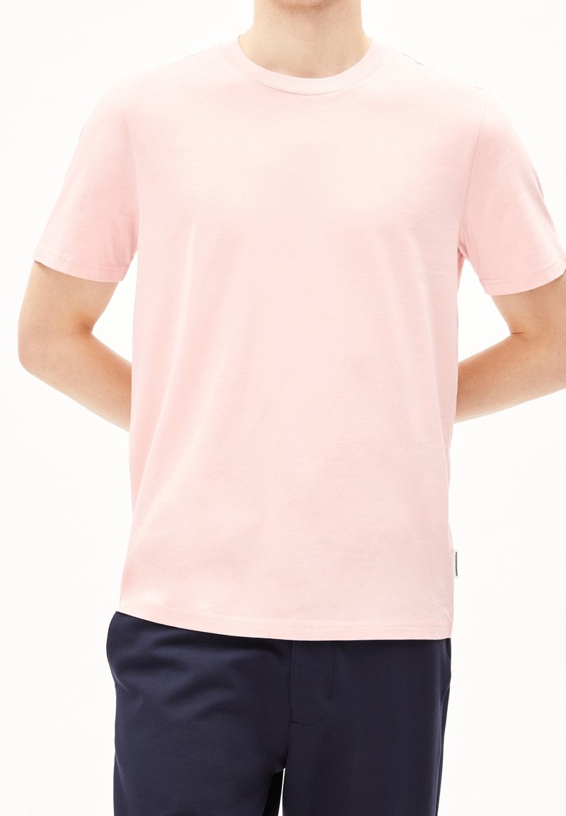Personne portant un t-shirt uni rose clair à manches courtes et un pantalon bleu marine foncé, debout les mains derrière le dos.