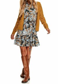 Šaty s květinovým vzorem a rozšířenou sukní, doplněné hořčicovým pleteným cardiganem. Hnědé kožené vysok�é boty ke kolenům dotvářejí outfit.