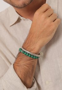 Braccialetto intrecciato in argento con perline a strisce verdi, indossato su un polso. Le perline variano in dimensione e sono lisce, complementando la texture metallica.