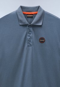 Šedé polo tričko vyrobené z měkké tkaniny, s třemi knoflíky, žebrovaným límečkem a kruhovou nášivkou s oranžovými akcenty.